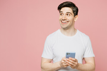 Young fun surprised man wearing white t-shirt casual clothes hold in hand using mobile cell phone look aside on area isolated on plain pastel light pink background studio portrait. Lifestyle concept.