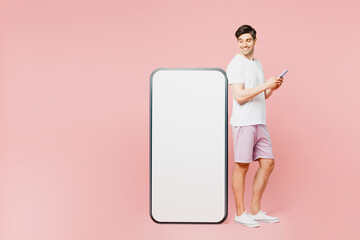 Full body side view young caucasian man wearing white t-shirt casual clothes look at big huge blank screen mobile cell phone with area using smartphone isolated on plain pastel light pink background.