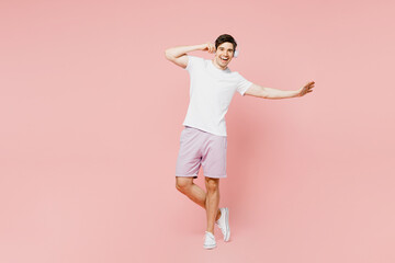 Full body smiling happy fun young caucasian man wearing white t-shirt casual clothes listening to music in headphones isolated on plain pastel light pink background studio portrait. Lifestyle concept.