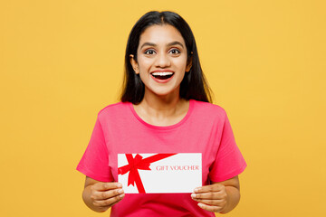 Young surprised fun happy Indian woman wearing pink t-shirt casual clothes hold gift certificate coupon voucher card for store isolated on plain yellow background studio portrait. Lifestyle concept.