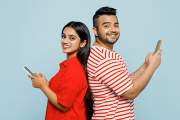 Young fun couple two friends family Indian man woman wear red casual clothes t-shirts together hold use mobile cell phone stand back to back look camera isolated on plain blue cyan color background.