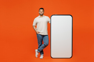 Full body fun young Indian man wear white t-shirt casual clothes big huge blank screen mobile cell phone smartphone with area looking camera isolated on plain orange red background. Lifestyle concept.