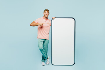 Full body young man wear pink t-shirt casual clothes point index finger on big huge blank screen mobile cell phone smartphone with workspace area isolated on plain blue background. Lifestyle concept.