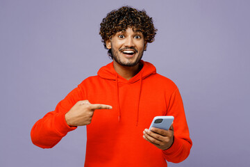 Young surprised fun Indian man wears red orange hoody casual clothes hold in hand use point index...