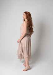 Full length portrait of beautiful  female model with long  brunette hair wearing a creamy pink gown dress. graceful dancing pose, with gestural hands  isolated on white studio background.
