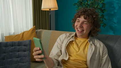 Young handsome man sitting on the sofa in the living room, watching a good movie and smiling widely