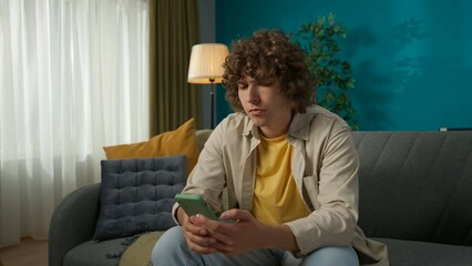Young man sitting on the sofa in the living room and scrolling through his smartphone, surfing in...