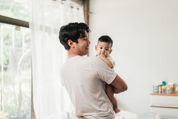 Happiness of fatherhood. Young Asian dad with adorable baby on his hands standing near window at home. Loving father spending time with infant child. Fathers day concept.