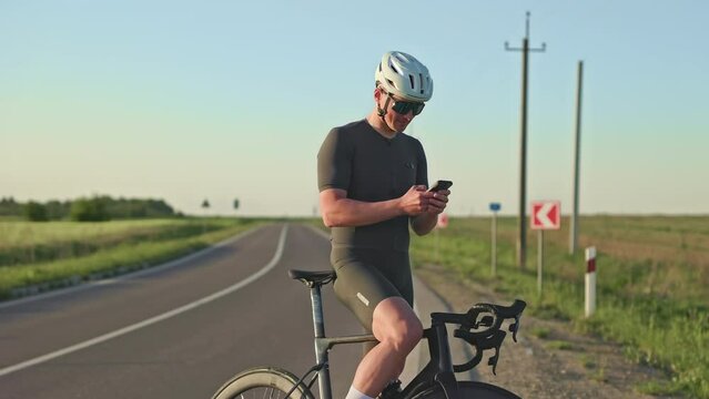 Well-built healthy man in sports clothing using smart device while holding black bike during short break on road. Delighted racer checking performance data via cycling computer app on phone.