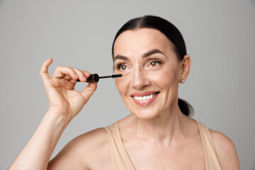 Close up of beautiful mature brunette woman smiling and applying mascara with makeup brush over grey background.