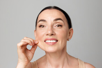 Close up beautiful mature woman pulling perfect skin and smiling a camera over grey background.