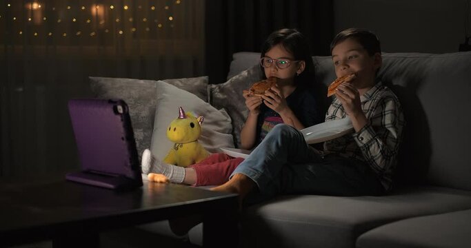 Children watching video on tablet on the sofa at home for entertainment or comedy. Excited kids siblings with tech for streaming a movie with online app in the living room while eating pizza
