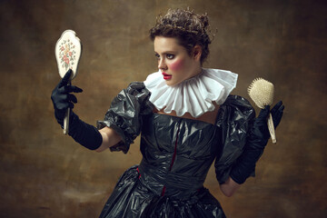 Portrait of woman in historic dress made of garbage bags, holding vintage hairbrush and looking at...