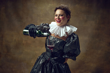 Portrait of cheerful woman in historic dress made of garbage bags, holding wine glass in hand and...