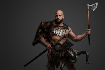 A fierce, muscular, bearded Viking warrior clad in furs and lightweight armor, with a helmet strapped to his waist, wielding two axes against a gray backdrop
