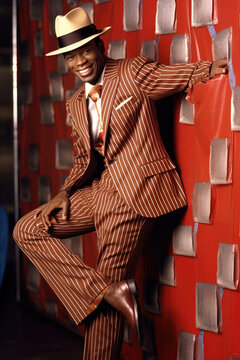 Confident African man in a stylish zoot suit and fedora, striking a relaxed pose with a bright white smile.