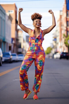 A tall African American woman with high-top fade haircut strikes a playful pose in a bright, patterned jumpsuit, large hoop earrings, and high heels.