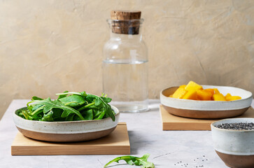Ingredients for green smoothie in ceramic bowls - greens, frozen mango, water, and chia seeds. Grey and yellow background, wooden stands