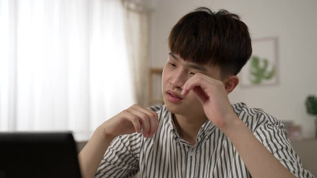 shoulder shot of a stressed asian man employee rubbing his sore eyes while working from home on the laptop in the living room at daytime.
