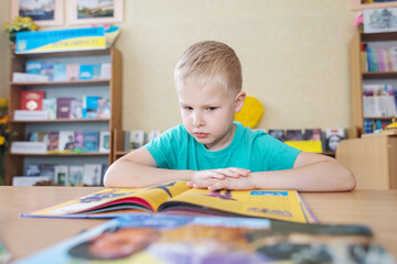 Fototapeta premium Boy in library. Seven year Ukrainian guy is sitting at a table in library and reading book. Concept of normal learning, thirst for knowledge, desire to learn new things. Blurred background and letters