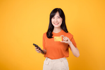 Fresh Asian woman in her 30s, using smartphone and presenting credit card on vibrant yellow...