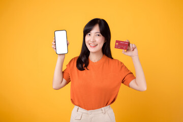 Naklejka premium Vibrant Asian woman in her 30s, showcasing smartphone and credit card on yellow background. Effortless online payment shopping.