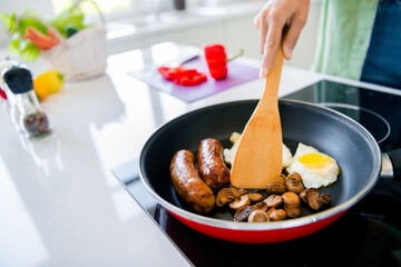 Cropped portrait of chef woman hand hold wooden spatula electric stove frying pan sausage mushroom egg homemade breakfast kitchen inside