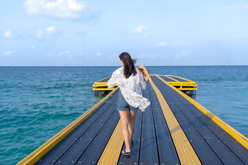 Back view of young Asian woman walking on a bridge looking back at the sea on a windy day Summer vacation. carefree concept Waves on background. Back view. Strong independent woman with flowing hair.