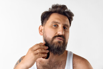 Obraz premium Handsome mature man in singlet taking care after beard with special brush against white studio background. Concept of men's beauty, skin care, cosmetology, health and wellness. Copy space for ad