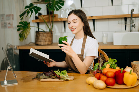 Young beautiful woman in the kitchen in an apron, fresh vegetables on the table, writes down her favorite recipes, comes up with ideas for dishes.