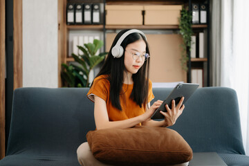 Obraz premium Attractive happy young Asian student studying at the college library, sitting at the sofa, using a laptop computer, tablet and headphones having a video chat.