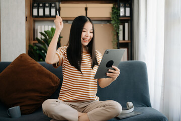 Obraz premium Attractive happy young Asian student studying at the college library, sitting at the sofa, using a laptop computer, tablet and headphones having a video chat.