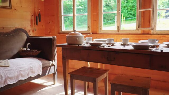 Rustic setting of wooden table and window at rural chalet, antique couch with white dinnerware. Humble ceramic plates, teacups, and utensils on aged table