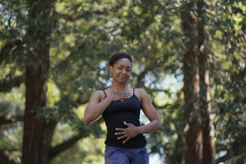 With one hand on their chest and one hand on their navel, a person stands tall in a forest, taking a break from yoga to pose for a portrait. They are standing with calm, certainty, poise and strength.