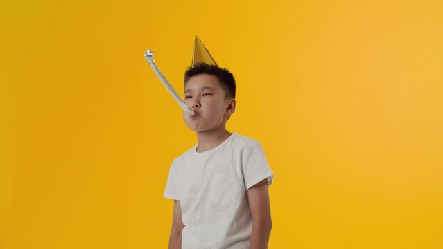 Young asian boy in a party hat blows in a party blower and crosses his hands on his chest with a dissatisfied face on a yellow background. Concept of boredom of a child on a holiday