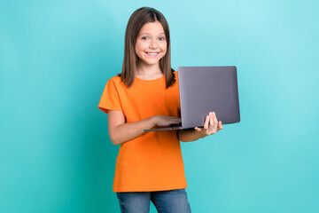 Photo of smart clever positive little girl wear orange stylish t-shirt holding laptop typing email isolated on teal color background