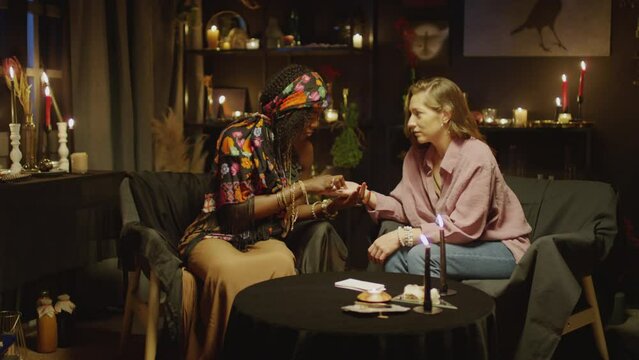 Medium shot of female fortune teller reading palm of woman explaining what to expect in near future while having seance in studio