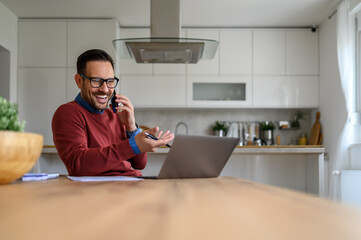 Cheerful male freelancer talking over smart phone and looking at laptop on desk in home office