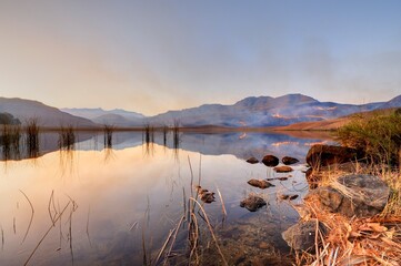 SMOKE FROM A WINTER GRASSFIRE reflects in a drakensberg mountain lake - 633340189