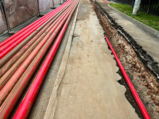 Red plastic pipes stacked on the pavement and one pipe in a trench