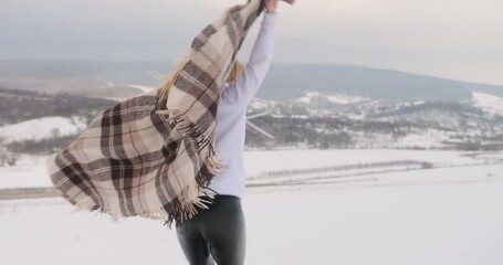 Young woman with blanket on shoulders having fun outside. Cheerful girl laughs and jumps in snow on winter mountains background