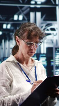 Vertical video Aged IT admin in modern server room holding clipboard during data center maintenance, crossing off completed tasks. Trained specialist in high tech workspace finishing checkup on