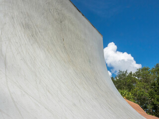 Low angle view of the steep, curved transition of a concrete vert ramp for experienced riders to perform aerial flips and spins in a public skate park, with partial view of trees, clouds, and sky