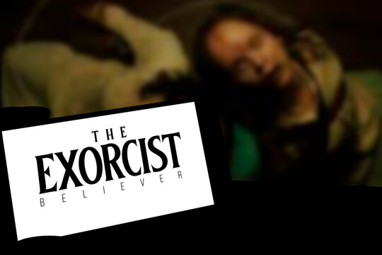 The Exorcist Believer logo is displayed on a smartphone screen with the actor from the movie in the background on the television screen.