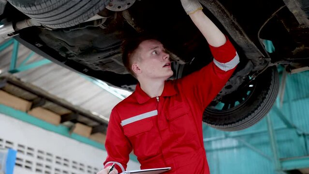 Caucasian mechanic man wearing safety red uniform inspect car suspension after accident, lifestyle multitasking small business Garage reparing service concept, technician examine damage automotive