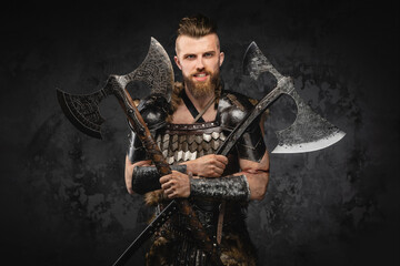 Fototapeta premium A menacing bearded viking warrior in fur and light armor showcasing his strength with two crossed axes, set against a textured grey wall
