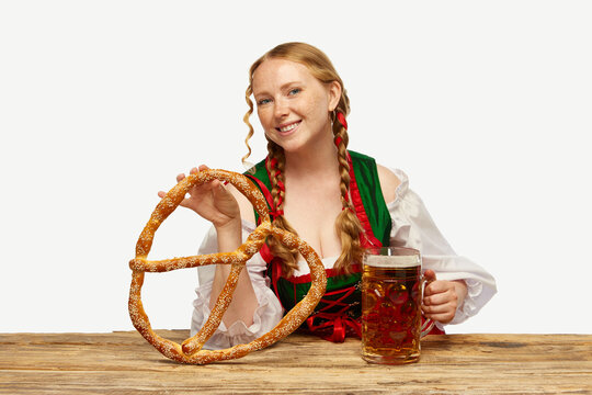 Young redhead girl waitress, wearing a traditional Bavarian or German dirndl, serving big beer mugs with huge pretzel. Oktoberfest concept