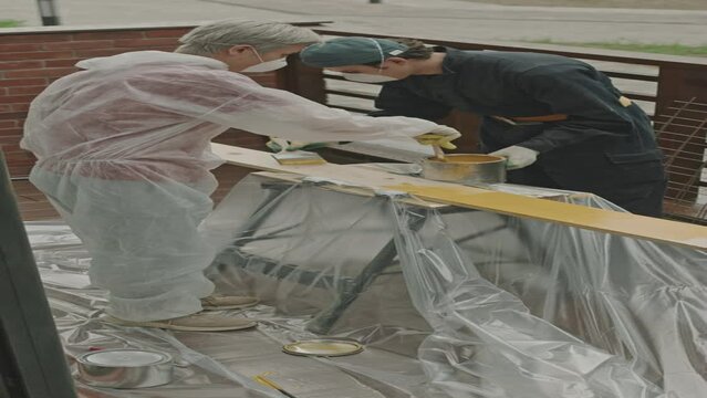 Vertical shot of two construction workers in respiratory masks and coveralls painting wooden plank on portable workbench standing on balcony of renovating apartment covered with polythene sheeting