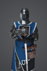 Medieval guard dressed in armor and blue surcoat holding a sword, standing in statue pose, against...