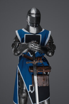 A medieval soldier garbed in blue surcoat and armor holding a sword with a still, statue-like pose, against a gray background